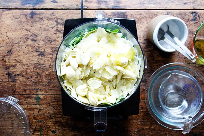 A food processor filled with basil, olive oil, salt, garlic, lemon, and parmesan.