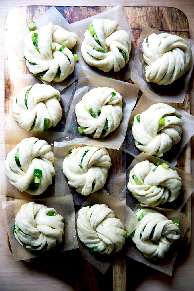 A board with 12 shaped Chinese scallion buns, each on a square of parchment paper.