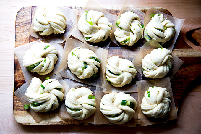 A board with 12 shaped Chinese scallion buns, each on a square of parchment paper.