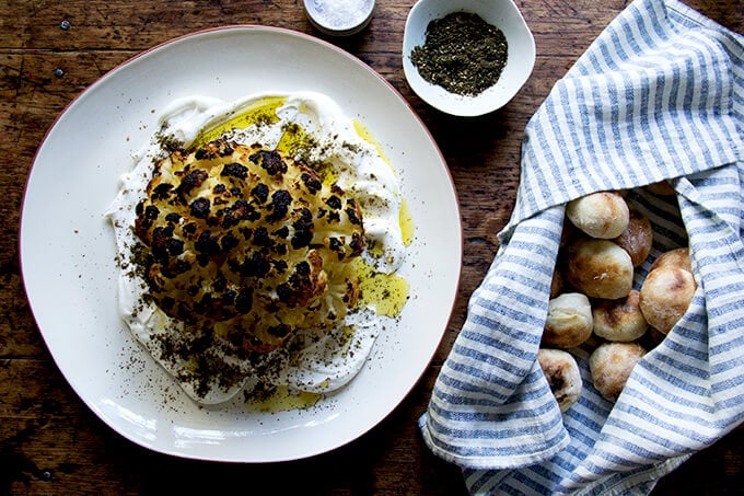 A platter topped with a head of whole roasted cauliflower aside a towel filled with homemade mini pita.