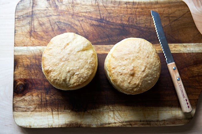 Two small loaves of bread on a board aside a serrated knife.