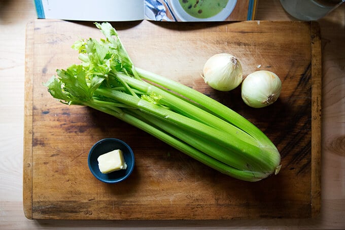 An overhead shot of a board with a head of celery on it aside onions and butter.