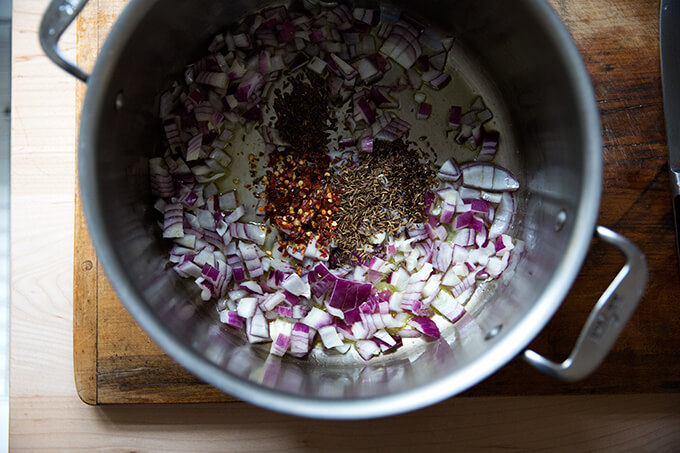 A pot with oil, diced red onion, cumin, and chili flakes. 