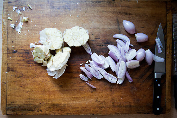 A cutting board with halved heads of garlic and sliced shallots. 