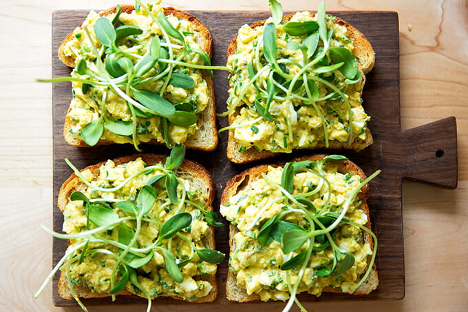 Four slices of toasted sourdough bread topped with avocado-egg salad and sprouts. 