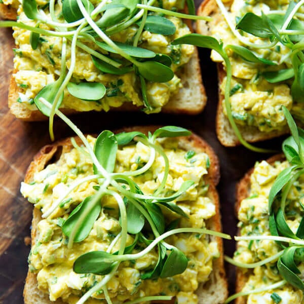 Four slices of toasted sourdough bread topped with avocado-egg salad and sprouts.