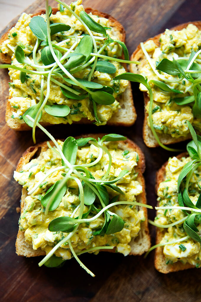 Four slices of toasted sourdough bread topped with avocado-egg salad and sprouts. 