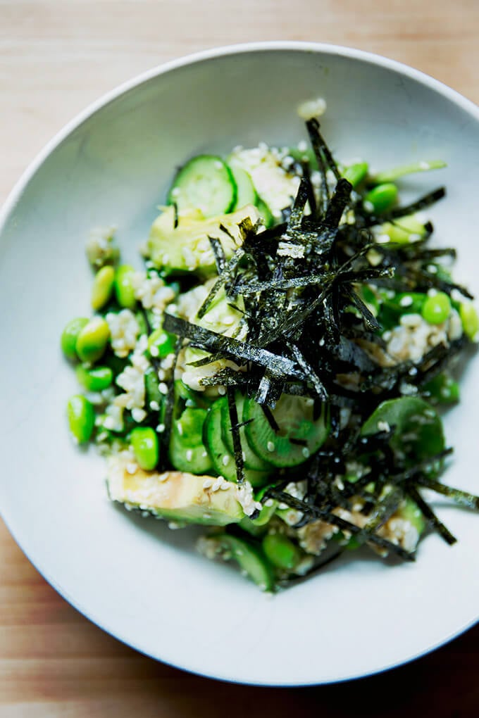 A bowl of sushi salad with brown rice, avocado, edamame, and cucumber with a sesame miso dressing, nori and sesame seeds.