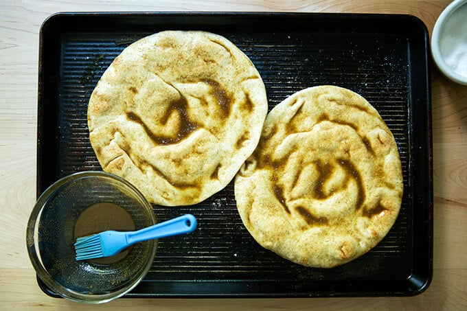 A sheet pan holding two rounds of pita brushed with oil, cumin and salt. 