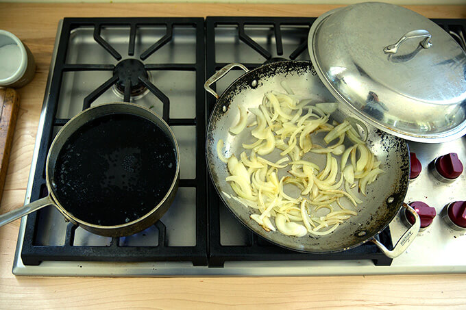 A stove top with a pot of black lentils simmering and a skillet with onions sauteing.