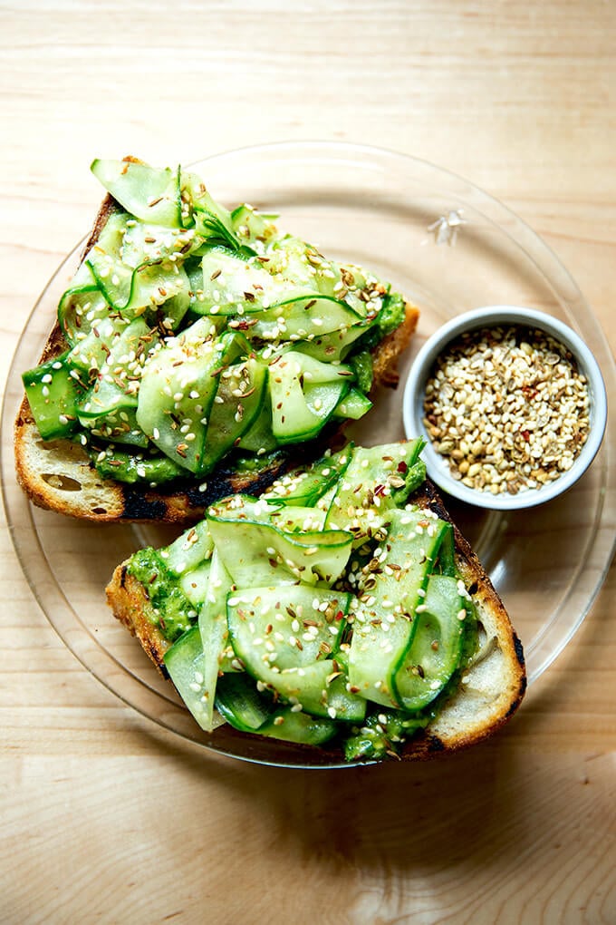 A plate with toast, green sauce, sliced cucumbers, and dukkah.