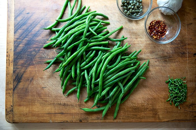 A cutting board with stemmed green beans, capers, garlic, and chili flakes.