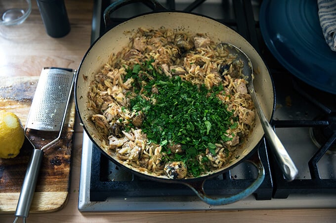 A skillet on the stovetop with chicken, orzo, artichokes, cheese, and, parsley