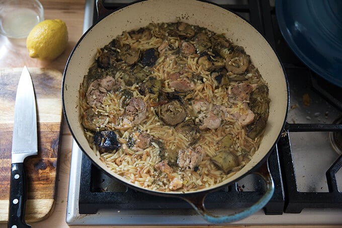 A skillet on the stovetop with chicken, orzo, and artichokes.