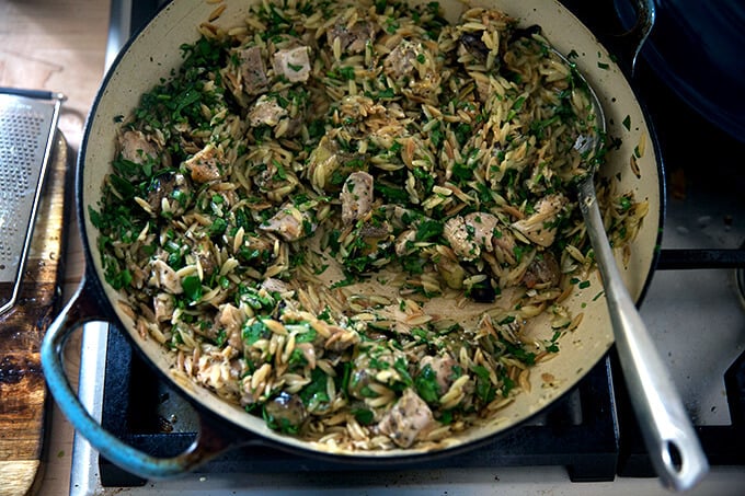 A skillet on the stovetop with chicken, orzo, artichokes, cheese, and, parsley all stirred together.