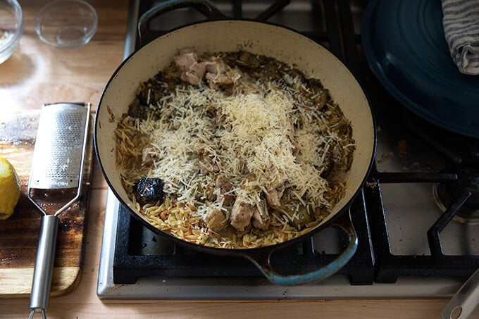 A skillet on the stovetop with chicken, orzo, artichokes, and parmesan cheese.