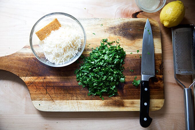 A board with a bowl of cheese and a heap of chopped parsley.