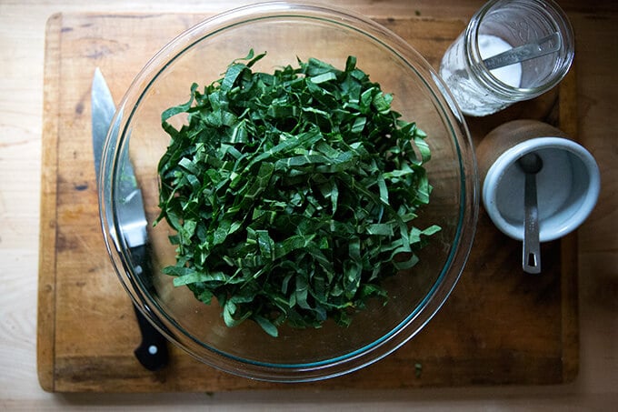 A bowl of raw, slivered collard greens aside a knife, and bowls of salt and sugar.