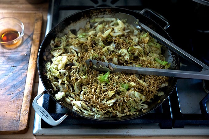 A large sauté pan filled with mushrooms, cabbage, ramen noodles, and scallions.
