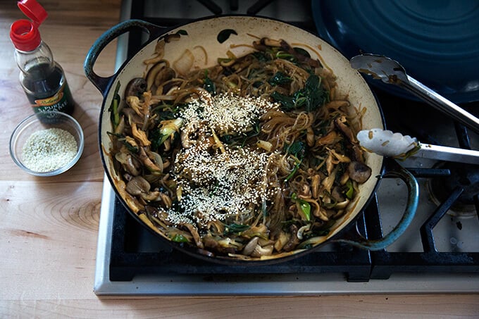 A braiser on the stovetop filled with japchae, sesame seeds, and sesame oil.