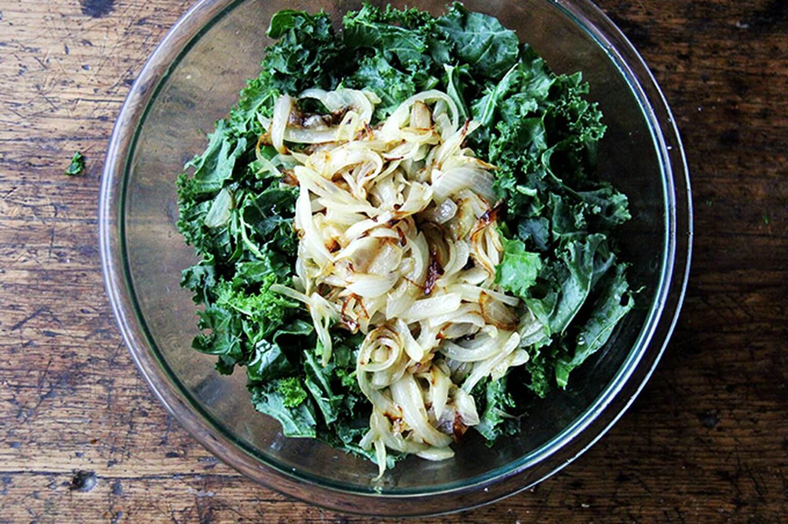 An overhead shot of a glass bowl filled with raw kale and caramelized onions.