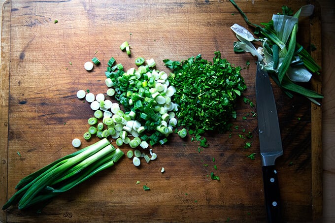 An overhead shot of a board filled with chopped scallions and parsley.