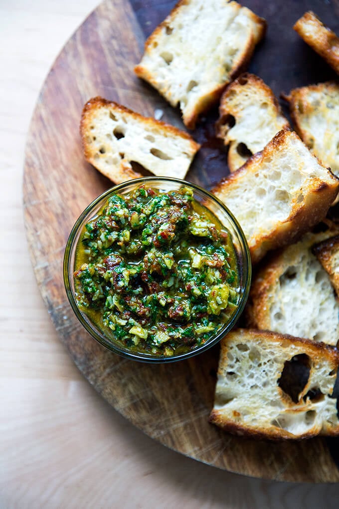 Sun-dried tomato and spinach pesto in a bowl aside toasty bread on a board.