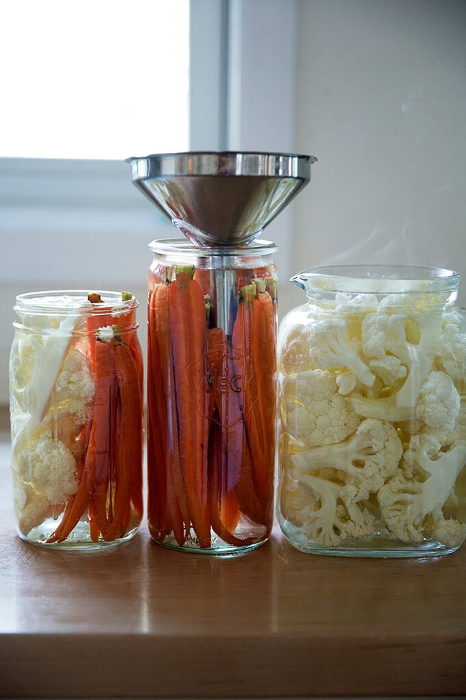 Pickled crudité in glass jars, one with a funnel.