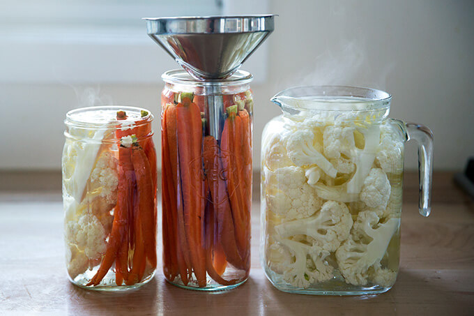 Pickled crudité in glass jars, one with a funnel.