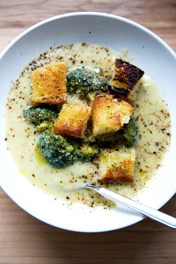 A bowl of broccoli-cheddar soup with olive oil toasted croutons and a spoon.