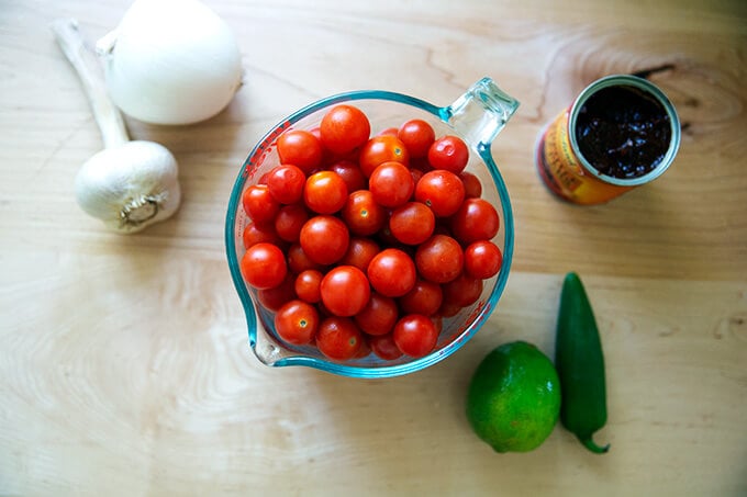 A bowl of tomatoes aside a head of garlic, an onion, a lime, a jalapeno and a chipotle in adobo sauce. 