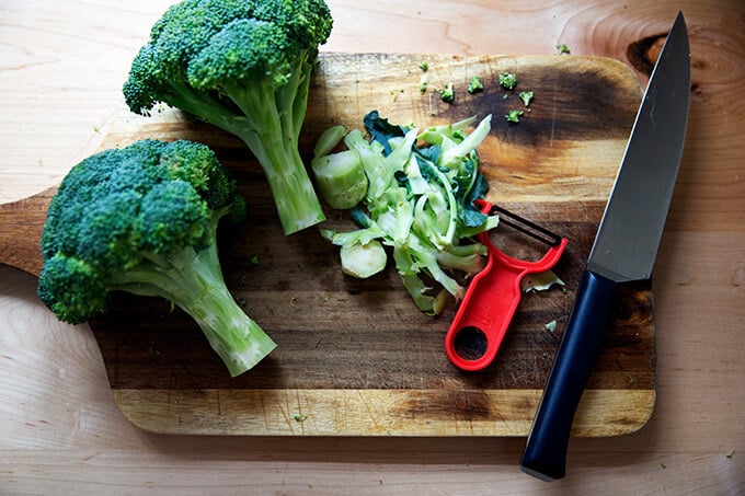 Two heads of broccoli on a cutting board, ends peeled with a peeler.
