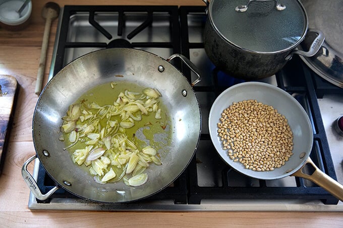 A stovetop with three different skillets: one holding shallots and garlic, one holding pine nuts, and one holding water.