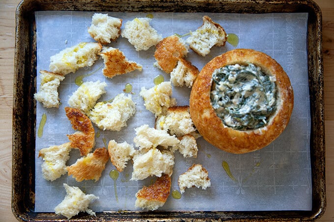 A sheet pan filled with a peasant bread bread bowl filled with spinach-artichoke dip.