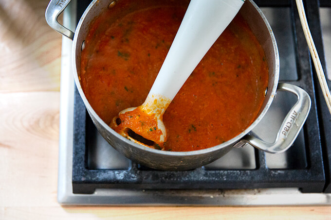 A pot of homemade tomato sauce simmering stovetop with an immersion blender inside.