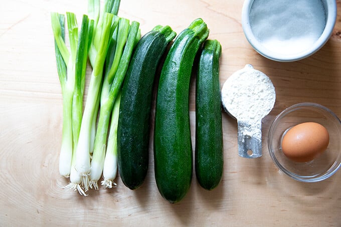 Scallions, zucchini, flour and an egg, the ingredients to make zucchini fritters, on a board. 