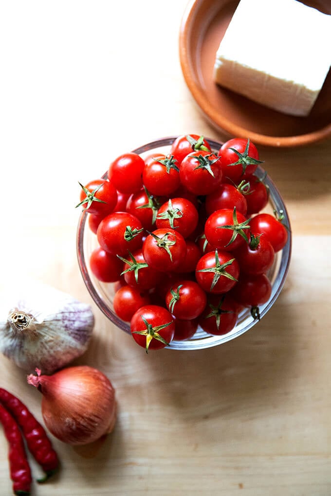 A bowl of cherry tomatoes aside a plate of feta and a head of garlic, shallots, and chilies.