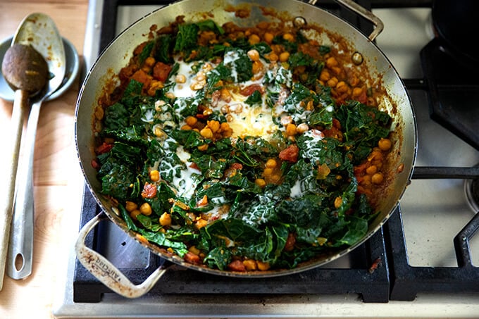 A skillet on the stovetop holding spicy chickpeas, tomatoes, and kale.