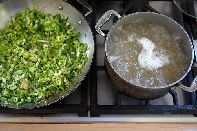 A pot of water boiling with pasta in it aside a skillet of sautéed leeks.
