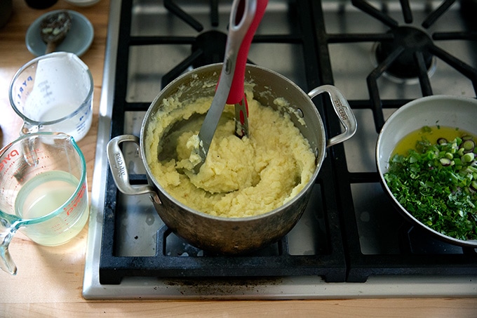A pot of mashed potatoes on the stovetop with a potato masher inside it.