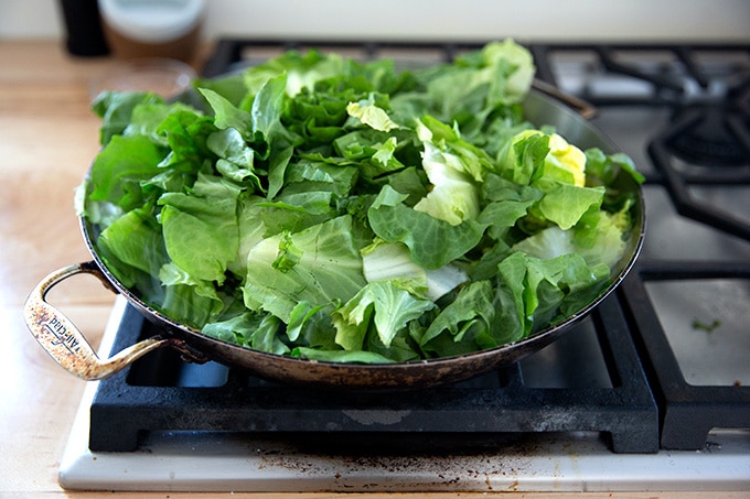 A large skillet stovetop with some olive oil, slice garlic, crushed red pepper flakes, and a heap of escarole inside.