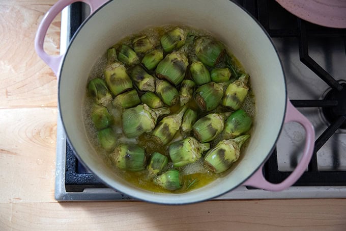 A large pot on the stovetop filled with baby artichokes cooking in olive oil, water, rosemary and garlic. - 5