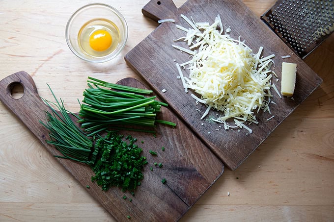 Two cutting boards holding chopped chives and grated Gruyere cheese aside a bowl holding an egg.