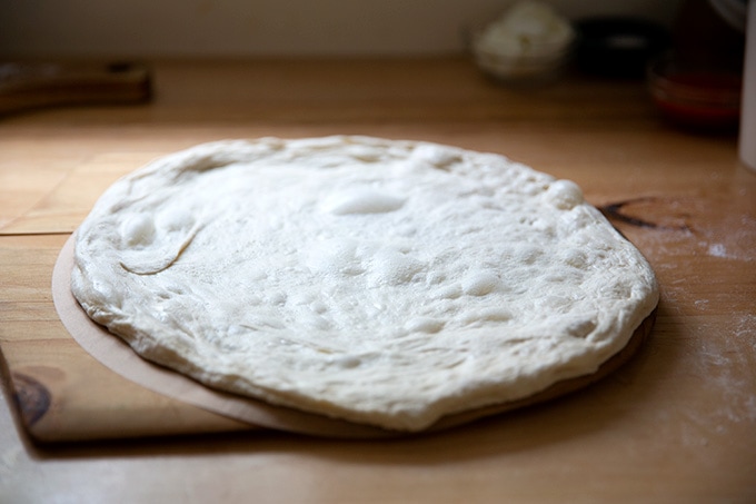 An unbaked round of pizza dough on a pizza peel lined with a parchment paper round.
