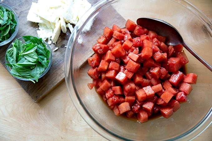 Cubed watermelon dressed with oil and vinegar aside a board with feta and herbs.