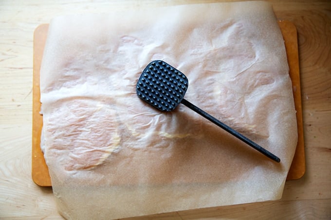 A mallet over a sheet of parchment paper over chicken breasts on a cutting board.