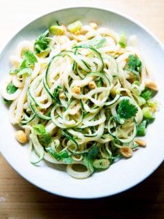 A bowl of peanut noodles with cucumbers, scallions and peanuts.