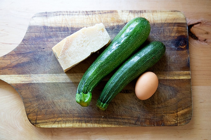 A block of parmesan, two zucchini, and an egg on a cutting board.
