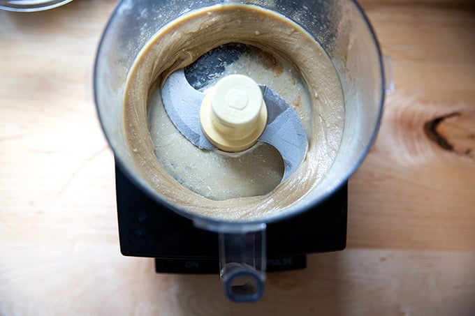 A food processor holding pureed tahini, salt, lemon, garlic, and cumin.