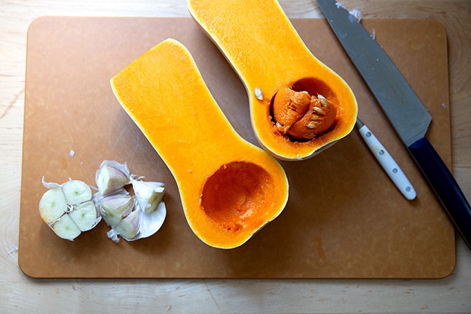 A halved butternut squash on a cutting board aside a halved head of garlic.